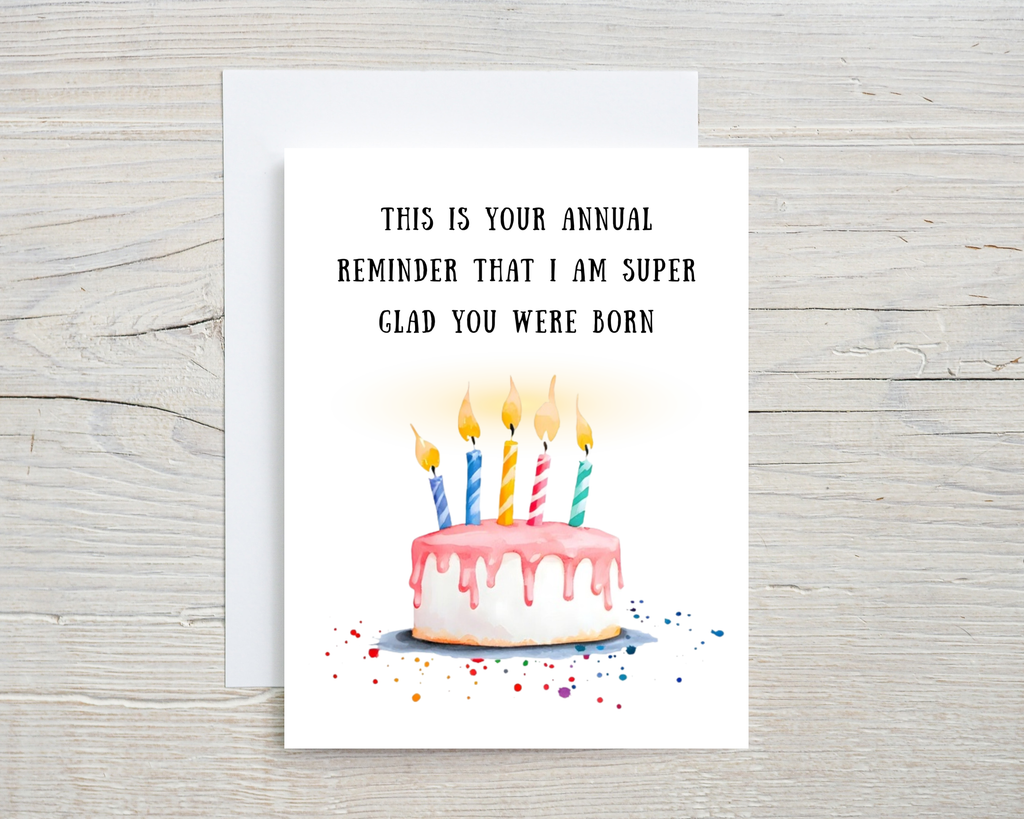 Birthday card with cake illustration and humorous text on a wooden surface "This is your annual reminder that I am super glad you were born"