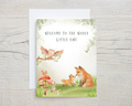 Greeting card with forest animals and 'Welcome to the World Little One' text on a wooden surface.